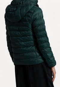 Veste en duvet vert foncé avec une capuche repliée, présentant une texture matelassée, une fermeture éclair et des poignets élastiques.