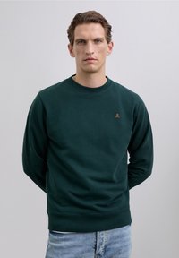 Homme portant un sweat-shirt vert foncé à col rond avec un petit logo de crâne orange et un jean bleu clair, se tenant avec les mains derrière le dos.