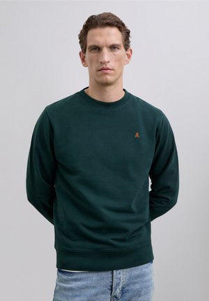 Man in een donkergroene sweatshirt met ronde hals en een klein oranje schedellogo, gekleed in lichtblauwe jeans, staand met zijn handen achter zijn rug.