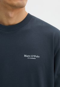 Marineblauer Pullover mit einem gerippten Rundhalsausschnitt. Auf der Vorderseite befindet sich ein weißes Logo mit der Aufschrift "Marc O'Polo EST. IN STOCKHOLM". Glatte Textur.