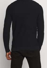 Svart stickad tröja med ett strukturerat mönster, med långa ärmar och ribbad nederkant. Bakifrån syns en figurnära design med minimal detaljering.