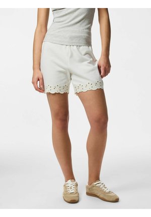 Žena v bielych krátkych šortkách so zvlnneným okrajom a kvetinovými výrezmi, sivom tielku bez rukávov a béžových teniskách stojí na bielom pozadí.