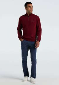 Bordeaux zip-up trui met een gebreide structuur en klein logo, gecombineerd met marineblauwe broek en witte sneakers.