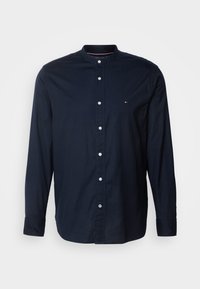 Chemise bleu foncé à manches longues avec boutons, col mandarin et petit logo sur la poitrine gauche sur fond uni.