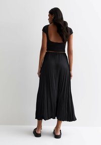 Top negro cortado con mangas en forma de capucha y espalda baja, combinado con una falda maxi negra plisada y fluida. Sandalias negras planas completan el look.