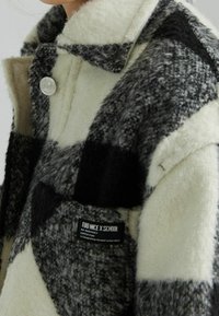 Fleecefodrad rutjacka i svart, vitt och grått. Har en spetsig krage, silverfärgad knappstängning och en märkesetikett framtill.