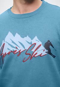 Hanorac turquoise cu grafice albe de munte. „Après Ski” în scris roșu, cu o siluetă de schior neagră. Material moale, dintr-un amestec de bumbac.