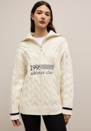 Jeune femme portant un pull en maille torsadée crème avec une fermeture éclair au quart et « 1995 athletics club » brodé sur le devant, associé à un pantalon noir.