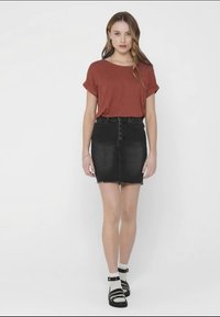 T-shirt corto marrone a maniche corte abbinato a una gonna di jeans nera con cinque bottoni. Il modello indossa calze fino al ginocchio con sandali neri.