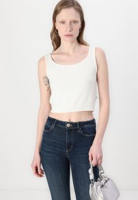 White cropped tank top with a round neckline, textured ribbing at the hem, paired with dark blue high-waisted jeans and a silver handbag.