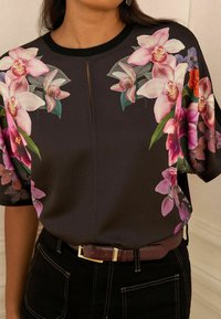 Blusa nera con grandi stampe floreali di orchidee rosa e viola sulle spalle e sulle maniche, abbinata a pantaloni scuri e una cintura marrone.