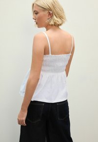 Witte gesmockte top met dunne bandjes, uitlopende onderrand en kanten accenten. Gepaard met donkere high-waisted jeans, gezien vanuit de achterkant.