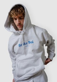 Helle graue Kapuzenjacke mit einer vorderen Tasche, die blauem Text "Broke Boi" und "POCKIES" bedruckt ist. Aus weichem Material mit einer großen Kapuze gefertigt.