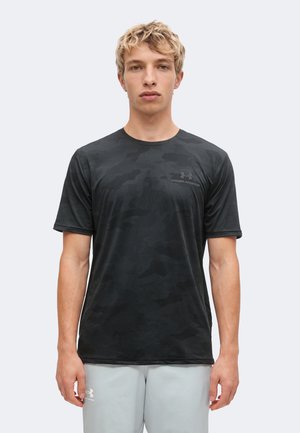 Jeune homme aux cheveux blonds courts portant un t-shirt noir à motif camouflage Under Armour et un short gris clair Under Armour, debout face à l'avant.