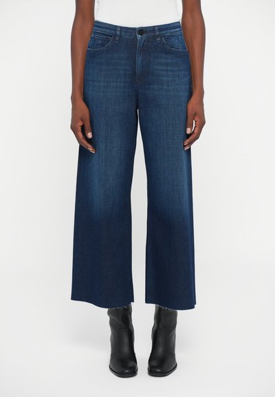 Persoon in hoge taille, wijde donkerblauwe jeans met ruwe randen en zwarte enkellaarsjes, staand tegen een witte achtergrond.