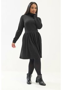 Czarna sukienka z długim rękawem, z wysokim kołnierzem i marszczonym wcięciem w talii, w połączeniu z czarnymi botkami na kostkę i rajstopami. Gładki materiał, dopasowana góra.