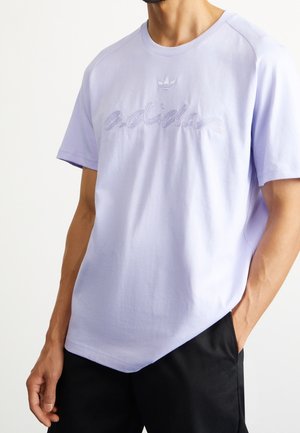 Man wearing light purple Adidas t-shirt with embroidered logo paired with black pants, standing with one hand in pocket.