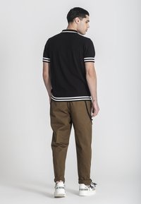 Černé polokošile s texturou a bílými pruhy na límci a rukávech, spárované s hnědými kalhotami a bílými teniskami s černými akcenty.