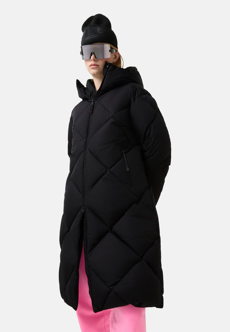 Zwarte pufferjas met een diamantvormig doorgestikt ontwerp, capuchon en zakken met rits. Draagt een zwarte beanie en reflecterende zonnebril.