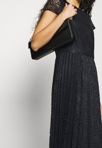 Robe en dentelle marine à manches courtes, au motif délicat, associée à une pochette en cuir noir avec quincaillerie dorée et un design épuré.