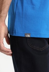 Blå t-shirt i bomull med en etikett märkt "The North Face" på den nedre vänstra sidan, kombinerad med mörka denimjeans. Närbild på tröjans fåll.
