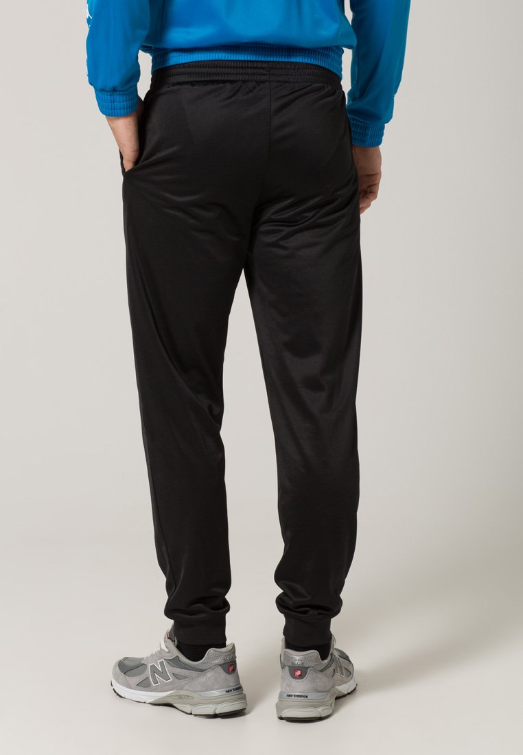 Uomo con pantaloni jogger neri, giacca blu e scarpe New Balance grigie in piedi con le mani nelle tasche su uno sfondo neutro.
