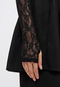 Μαύρο ένδυμα με υφασμάτινη υφή και μακριά μανίκια από λουλουδάτη δαντέλα. Κοντινό πλάνο ενός χεριού που φοράει ένα απλό ασημένιο δαχτυλίδι.