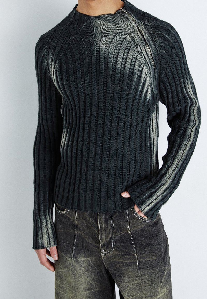 Mörkgrå ribbad stickad tröja med hög hals och långa ärmar, med en subtil gradienteffekt och synliga sömmar. Avslappnad passform.