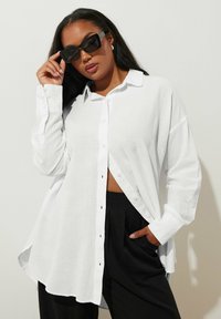 Witte blouse met lange mouwen, knoopsluiting en een ontspannen pasvorm, met zijsplitten en een gestructureerde stof. Gecombineerd met zwarte high-waisted broek en een zonnebril.