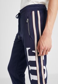 Marina joggers i mjukt tyg med en vit logotyp, sidfickor med dragkedja och röda och krämfärgade ränder längs de yttre sömmarna.