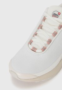 Fehér sneaker textúrált hálós felsőrésszel, rózsaszín díszítéssel a fűzőn, és kiemelkedő párnázott talppal. A nyelvén egy kör alakú logó található.