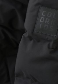 Svart vadderad jacka med en strukturerad, matt yta och en rektangulär svart lapp märkt "COLOR KIDS." Detaljerad sömnad och jämn yta synlig.