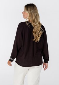 Femme aux longs cheveux blonds ondulés portant un pull texturé sombre et un pantalon blanc, se tenant de dos avec la tête de profil.
