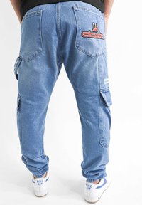 Hellblaue Denim-Cargohosen mit elastischen Bündchen, Seitentaschen und gesticktem Logo auf der Gesäßtasche. Weiße Sneakers mit blauen Akzenten.