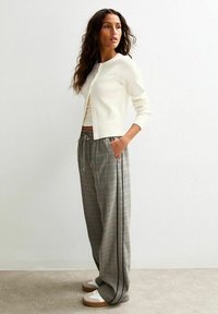 Krämfärgad ribbad cardigan över en ljus topp, kombinerad med lösa rutiga byxor i grått med gula accenter och svarta sidolinjer, avslutat med vita sneakers.