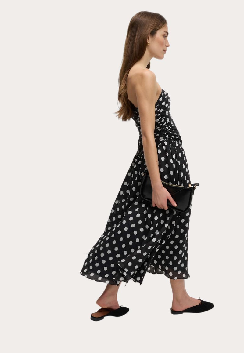 Abito nero a pois con corpino aderente, gonna fluida e dettagli arricciati. Indossato con una pochette nera e scarpe slip-on.