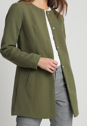 Vrouw draagt een open olijfgroene jas met knopen, wit topje en grijze geruite broek, met haar hand vlak bij de jaszak.