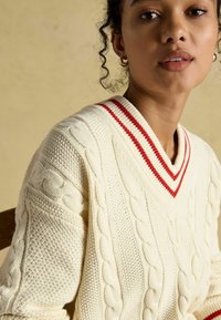 Kreemi värvi kudum, millel on tekstuurilised mustrid, V-kujuline kaelus ja punased triibud krae ääres.