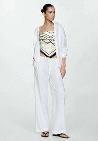 Pantaloni cropped bianchi in lino abbinati a un top beige senza spalline con motivi geometrici neri e una camicia bianca aperta.