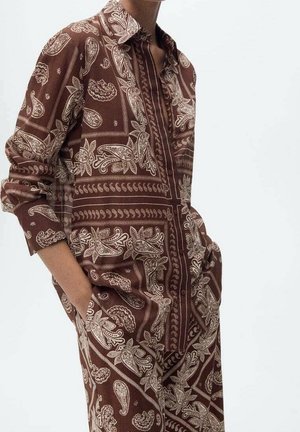 Osoba ubrana w brązową, wzorzystą paisley koszulę z długim rękawem i dopasowane spodnie, z rękami w kieszeniach, na gładkim tle.