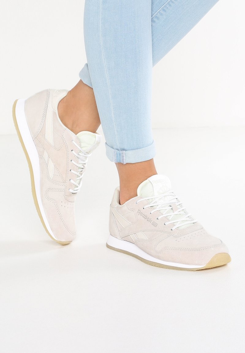 Ljusbeige mockaskor från Reebok med vita snören och sålar, bärs tillsammans med uppvikta ljusblå jeans mot en vit bakgrund.