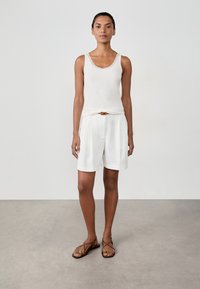 Donna in piedi con espressione neutra, indossa un top senza maniche bianco, pantaloni corti bianchi con cintura e sandali piatti marroni, su un pavimento grigio.