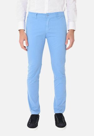 Uomo che indossa pantaloni slim fit azzurri, camicia bianca e sneakers nere con suola bianca, in piedi su sfondo bianco.