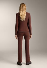 Hnedý priliehavý oblek s dlhými rukávmi a nohavicami so širokým strihom. Vyrobený z textúrovanej tkaniny, s čistým, moderným dizajnom a minimálnymi švom.