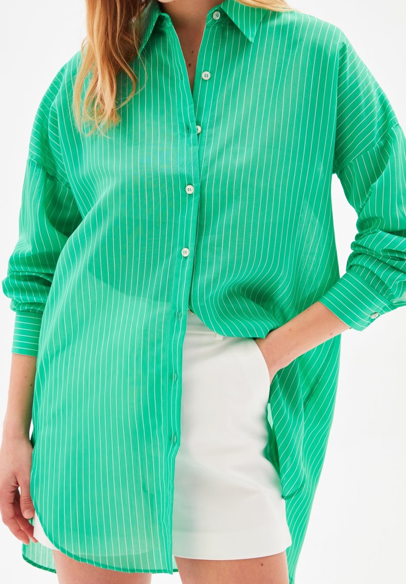 Femme portant une longue chemise verte à boutons avec des rayures blanches par-dessus une jupe blanche, une main dans la poche de la jupe, debout sur fond blanc.