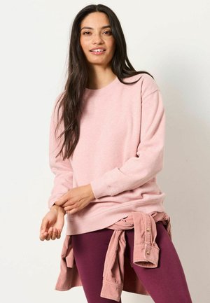 Frau mit langen dunklen Haaren, die einen hellrosa Pullover und dunkelrosa Hose trägt, mit einem rosa Hemd um die Taille gebunden, lächelt in die Kamera.