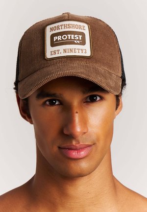Jonge man met een gemiddelde huidskleur die een bruine corduroy pet draagt met de tekst "Northshore Protest Est. Ninety3" op het voorste embleem.