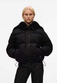 Zwarte pufferjack met een nepbontkraag, textuurstof, sluiting met knopen aan de voorkant en zijzakken. Bijpassende gestreepte broek.