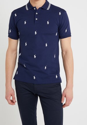 Mann trägt ein dunkelblaues Poloshirt mit weißen bestickten Polo-Spieler-Logos und passende dunkelblaue Hosen, steht vor einem schlichten Hintergrund.