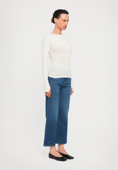 White long-sleeve top with a round neckline, paired with high-waisted dark blue wide-leg jeans and black flat shoes.